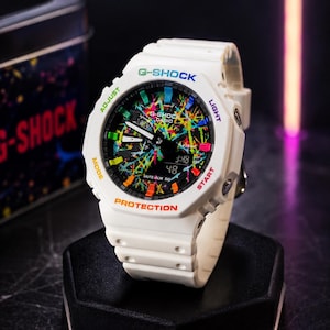 Könnte beinhalten: Eine weiße G-Shock-Uhr mit schwarzem Zifferblatt und einem bunten Farbspritzer-Muster. Die Lünette trägt die Aufschriften G-Shock, Adjust, Light, Mode, Start und Protection in verschiedenen Farben. Sie verfügt über analoge Zeiger und ein Display.