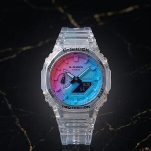 May include: A clear G-Shock Casio watch with a gradient dial of pink, purple, and blue. The watch face displays the time 10:08 and seconds 55. The watch has a clear band and is set against a dark background.