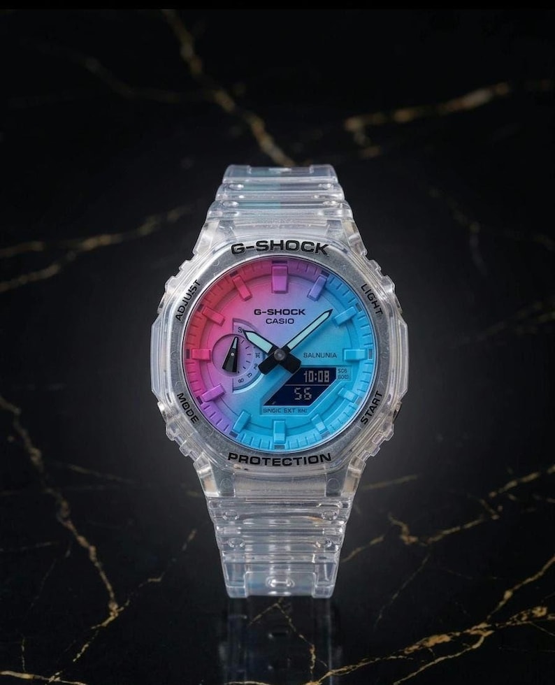May include: A clear G-Shock Casio watch with a gradient dial of pink, purple, and blue. The watch face displays the time 10:08 and seconds 55. The watch has a clear band and is set against a dark background.