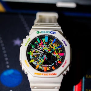 May include: White G-Shock watch with a colorful splatter design on the face. The watch features a digital display showing the time. The watch has the text "G-SHOCK" and "PROTECTION" on the face.
