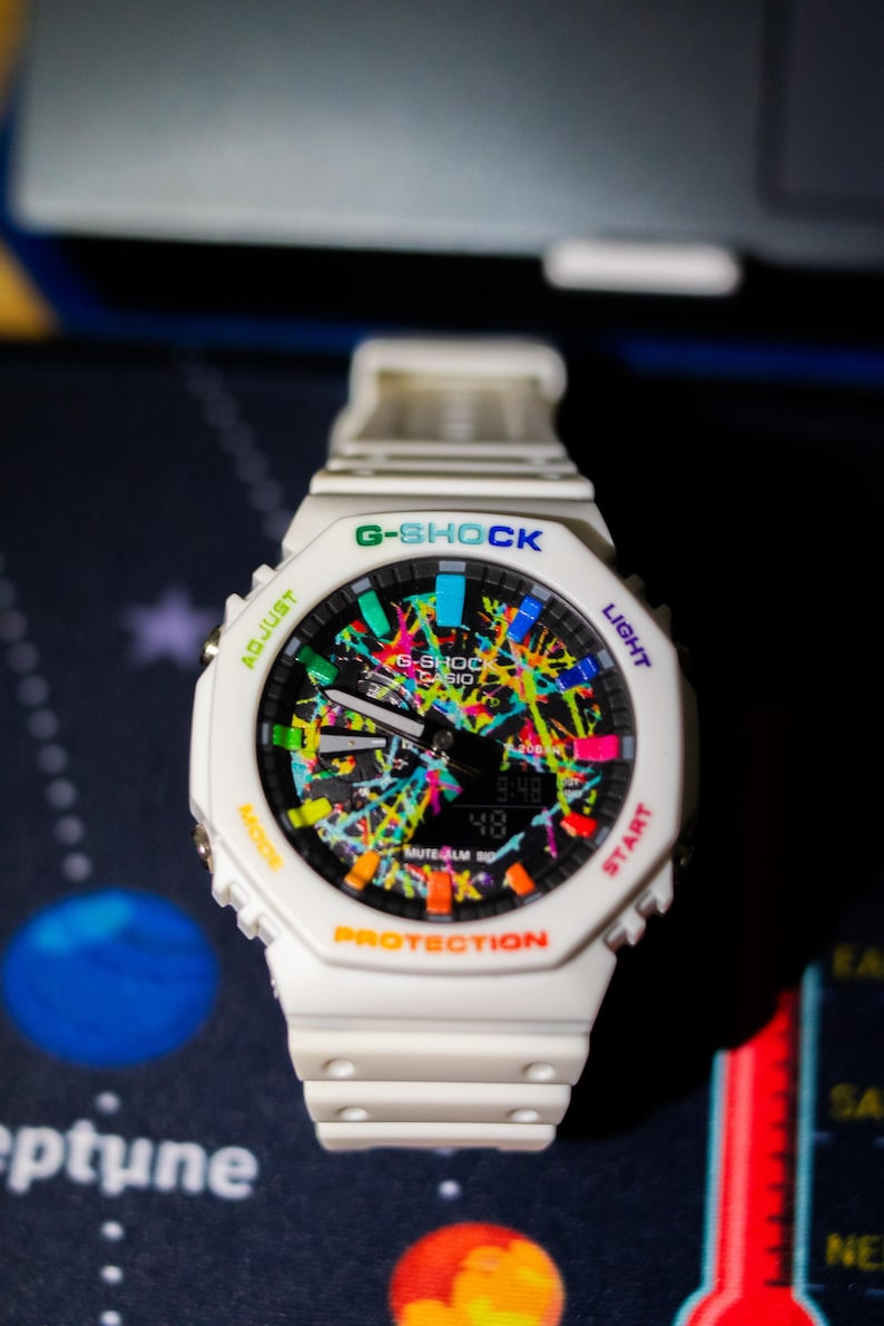 May include: White G-Shock watch with a colorful splatter design on the face. The watch features a digital display showing the time. The watch has the text "G-SHOCK" and "PROTECTION" on the face.