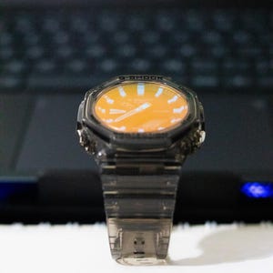 May include: A transparent, gray-tinted wristwatch with an orange face. The watch has a digital display and a clear band. The watch is set against a blurred background.