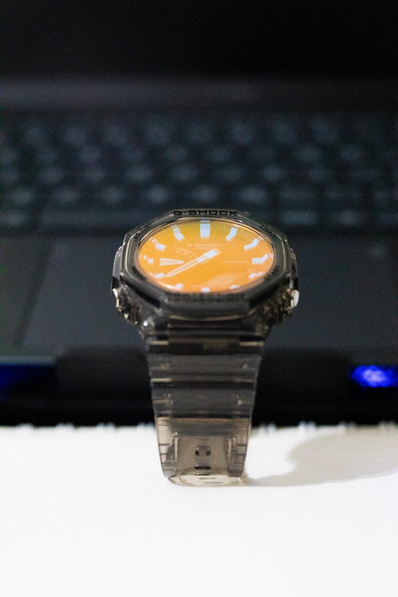 May include: A transparent, gray-tinted wristwatch with an orange face. The watch has a digital display and a clear band. The watch is set against a blurred background.