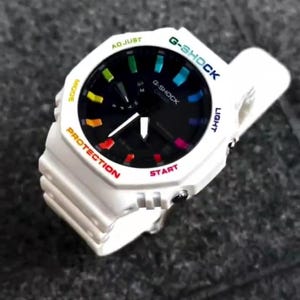 May include: White G-Shock watch with a black face and rainbow accents. The watch features a white band and bezel with text in various colors, including "G-SHOCK", "PROTECTION", and "START". The watch has a digital display.