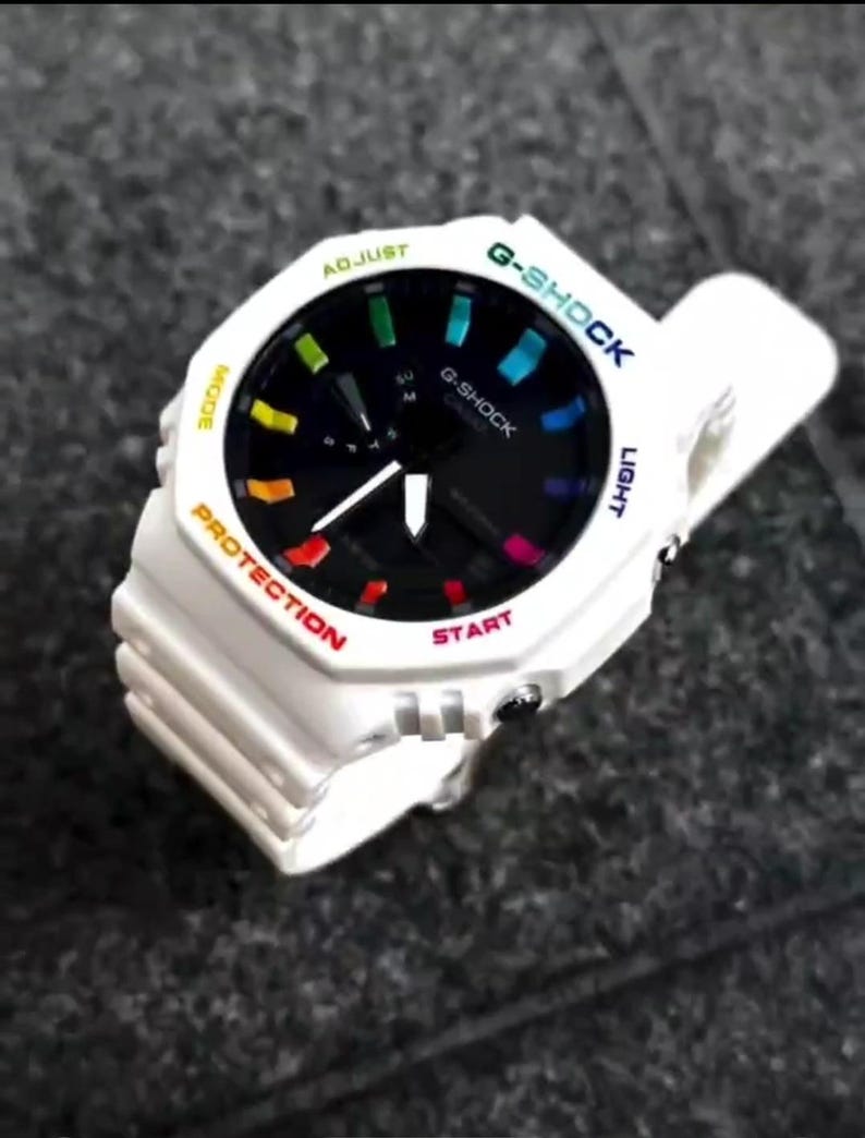 May include: White G-Shock watch with a black face and rainbow accents. The watch features a white band and bezel with text in various colors, including "G-SHOCK", "PROTECTION", and "START". The watch has a digital display.