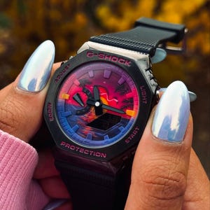 May include: Black G-Shock watch with a colorful sunset design on the face. The watch has a black band and a digital display showing the time. The watch is held by a person wearing a pink sweater.