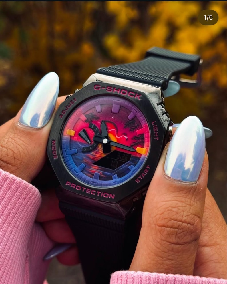 May include: Black G-Shock watch with a colorful sunset design on the face. The watch has a black band and a digital display showing the time. The watch is held by a person wearing a pink sweater.