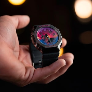 May include: A G-Shock watch with a black band and a colorful face featuring a sunset design. The watch has a silver-tone case and is held in a hand against a dark background. The watch face displays the words "G-SHOCK" and "PROTECTION".