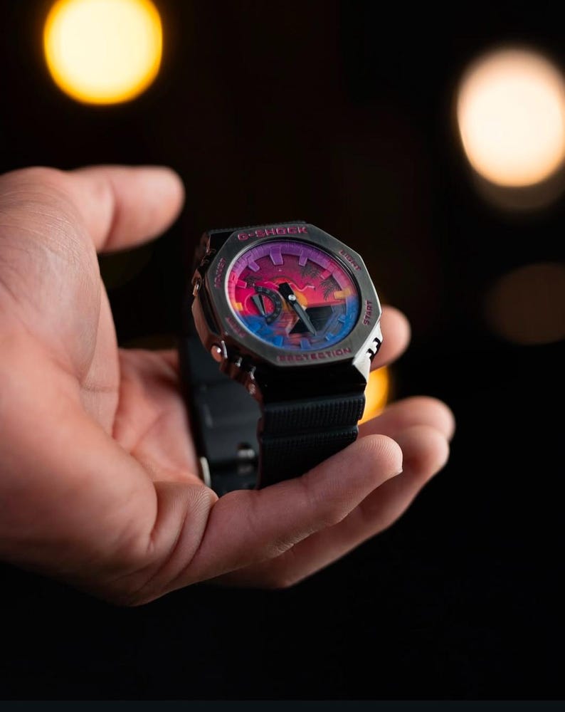 May include: A G-Shock watch with a black band and a colorful face featuring a sunset design. The watch has a silver-tone case and is held in a hand against a dark background. The watch face displays the words "G-SHOCK" and "PROTECTION".