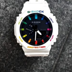 May include: White G-Shock watch with a black dial and colorful accents. The watch face displays the G-Shock and Casio logos, along with the text "ADJUST", "MODE", "LIGHT", "START", and "PROTECTION". The watch has a digital display and a white strap.
