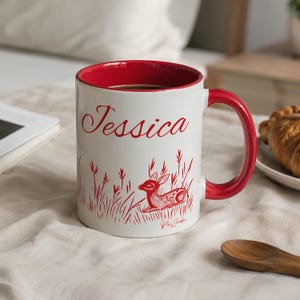 May include: A white ceramic mug with a red interior and handle. The exterior features the name Jessica in red script above a red illustration of a fawn resting in tall grass. The text Rose Zombie is printed in small red script below the fawn.