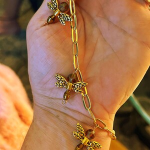 May include: Gold-toned chain bracelet featuring three dragonfly charms. Each dragonfly has detailed wings and a brown bead body. The bracelet's chain links are oval-shaped, and the overall design is elegant and nature-inspired.