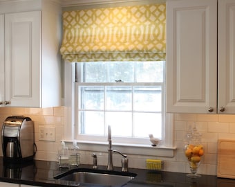 May include: A white kitchen with white cabinets, a black countertop, and a stainless steel sink. The window has a yellow and white patterned roman shade.