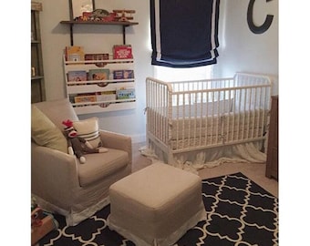 May include: A nursery with a white crib, a beige armchair, and a matching ottoman. A black and white patterned rug covers the floor. A wall-mounted bookshelf holds children's books. A dark blue window shade and a large letter "C" decorate the wall.