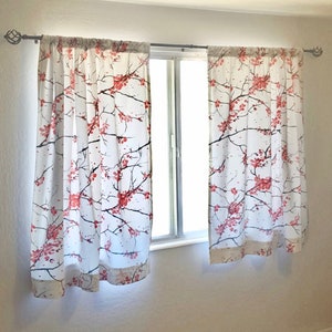 May include: White sheer curtains with a floral pattern of pink cherry blossoms on a white background. The curtains are hung on a window with a white trim.