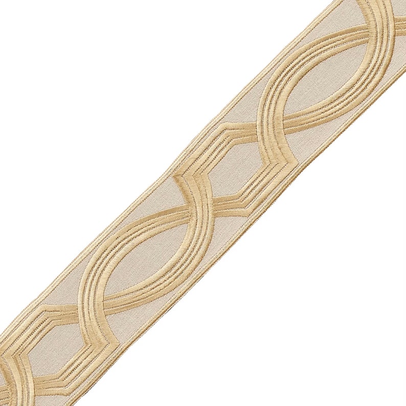 May include: Gold trim with a geometric pattern on a beige fabric. The trim has a repeating pattern of interlocking circles and squares.