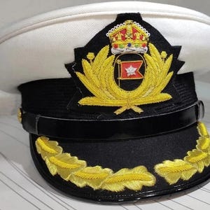 Könnte beinhalten: Eine weiß-schwarze Kapitänsmütze mit goldgestickten Details. Die Mütze hat eine schwarze Krempe mit goldenen Lorbeerblättern, ein schwarzes Band und eine weiße Oberseite. Auf der Vorderseite befindet sich ein goldenes Emblem mit Krone und rot-weißer Flagge.