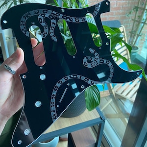 May include: A glossy black guitar pickguard featuring a detailed, etched silver-toned snake design that winds across the surface. The pickguard includes pre-cut openings for pickups, a bridge, and control knobs, along with several small screw holes.