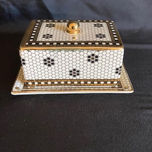 May include: A square ceramic trinket box with a matching tray. The box and tray feature a black and white honeycomb pattern with black floral accents. The box has a gold-colored rim and a gold knob on the lid.