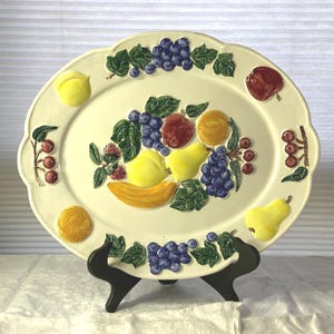 May include: Off-white oval serving platter with a raised fruit design. The platter features grapes, apples, pears, bananas, and other fruits in shades of red, yellow, and purple. The rim has a scalloped edge.