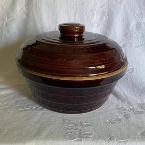 May include: A brown ceramic casserole dish with a lid. The dish has a round shape with a ridged design and a handle on the lid. The dish is sitting on a white tablecloth.