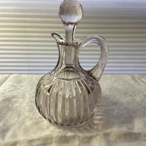 May include: A clear glass cruet with a round body and a handle. The cruet has vertical ribbed detailing and a faceted stopper. The cruet is designed for serving oil or vinegar.