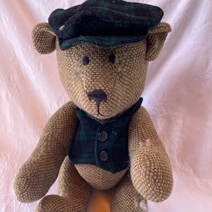 May include: A handmade teddy bear with a brown tweed body, wearing a dark green plaid vest and cap. The bear has a black nose and button eyes. The bear is a classic toy.