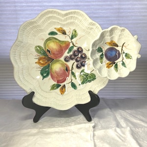 May include: Cream-colored ceramic serving dish with a scalloped edge and attached dip bowl. The dish is decorated with hand-painted pears, grapes, and leaves. The dish is supported by a black stand.