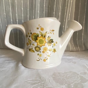 May include: A white ceramic watering can with a floral design in yellow, green, and brown. The watering can has a handle and a spout, and is sitting on a white surface.