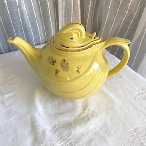 May include: A yellow ceramic teapot with a lid, spout, and handle. The teapot has gold trim and a decorative pattern of gold leaves. The teapot is sitting on a white surface.