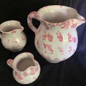 May include: A white ceramic pitcher and two matching creamers with a pink speckled pattern. The pitcher has a large handle and a pouring spout. The creamers have small handles. The set is likely for serving beverages.
