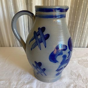 May include: A gray stoneware pitcher featuring a glossy finish, a curved handle, and hand-painted cobalt blue decorative patterns including cross-hatch marks and floral motifs, with blue bands encircling the rim and base.