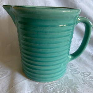 May include: A turquoise ceramic pitcher with a handle and a spout. The pitcher has a ribbed design around the body. The handle is curved and attached to the side of the pitcher. The pitcher is sitting on a white surface.