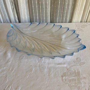 May include: A clear glass serving dish shaped like a large leaf with a pointed tip and serrated edges. The glass features a subtle blue tint along the outer rim and veins that radiate from the center stem, creating a textured surface.