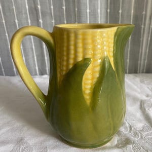 May include: A ceramic pitcher shaped like a corn cob with a yellow kernel texture on the upper portion and green husks wrapping around the base. The handle is a smooth, light green color, and the rim features a spout for pouring liquids.
