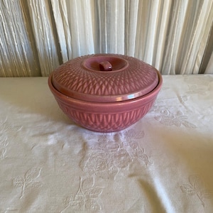 May include: A round, dusty rose-colored ceramic casserole dish with a matching lid. The exterior of both the bowl and the lid features a textured, repeating diamond-shaped pattern. A small, centered knob handle sits atop the lid.