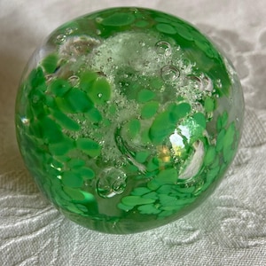 May include: A clear glass paperweight with a spherical shape. The interior features vibrant green leaf-like inclusions and air bubbles, creating a decorative effect. The paperweight is a unique decorative item.