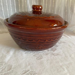 May include: A brown ceramic casserole dish with a lid. The dish has a round shape with a decorative pattern around the rim and on the lid. The lid has a small handle on top.