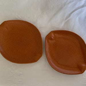 May include: Two brown, organically shaped plates with a speckled texture. One plate has a slight indentation on one side. The plates are set against a white, textured background.