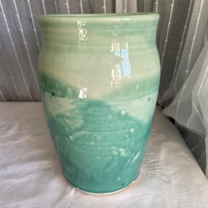 May include: A handmade ceramic vase with a cylindrical shape. The vase features a gradient of colors, transitioning from a light green at the top to a deeper turquoise at the bottom. The surface has a textured, speckled appearance.