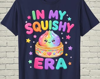 May include: A navy blue t-shirt featuring a colorful graphic of a rainbow-colored dumpling with a smiling face sitting in a bamboo steamer. The text "IN MY SQUISHY ERA" is printed in large, multi-colored bubble letters, surrounded by small stars.