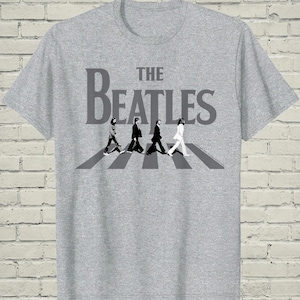 May include: Gray t-shirt featuring the band name "The Beatles" in large, gray letters above a black and white image of the band members walking across a crosswalk. The shirt is made of a soft material.