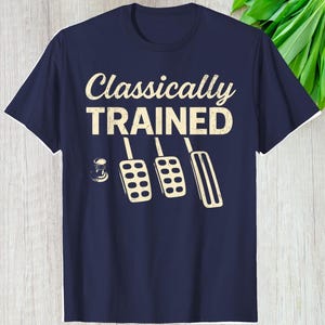 May include: Navy blue t-shirt with the text "Classically TRAINED" in a distressed font. Below the text are illustrations of a gear shift, clutch, brake, and accelerator pedals in a vintage style.