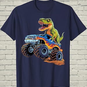 May include: A navy blue t-shirt featuring a graphic print of a green Tyrannosaurus Rex standing on top of a blue monster truck. The truck has large black tires and orange and yellow flame decals on the side, depicted driving over brown dirt.