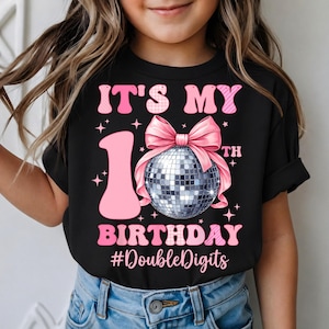 May include: Black t-shirt with pink text and graphics. The text reads "IT'S MY 1ST BIRTHDAY #DoubleDigits". A pink bow and disco ball are featured in the design. The shirt is worn by a person.