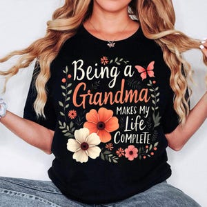 May include: Black t-shirt with the text "Being a Grandma Makes My Life Complete" in white and orange script. The design includes floral and butterfly accents in orange, pink, and white.