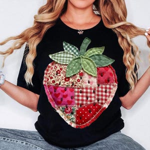 May include: Black t-shirt featuring a large, patchwork strawberry design. The strawberry is composed of various fabric patterns, including floral, heart, and gingham prints, with green leaves and stem. The shirt is a casual, everyday wear item.
