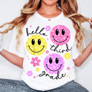 May include: A white t-shirt featuring three smiley faces in yellow, pink, and light purple, each patterned with small white flowers. The shirt includes the black cursive text hello third grade, along with small pink and yellow flower and star icons.