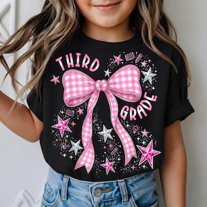 Puede incluir: Una camiseta negra con un gran gráfico de lazo a cuadros vichy rosa y blanco en el centro. El texto "THIRD GRADE" está impreso en rosa alrededor del lazo. El diseño incluye estrellas rosas y plateadas, libros, lápices y bombillas.