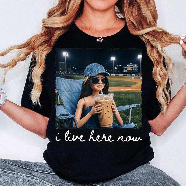 K&ouml;nnte beinhalten: Schwarzes T-Shirt mit einer Grafik einer Puppe mit Sonnenbrille und Jeansm&uuml;tze, die einen Eiskaffee h&auml;lt. Die Puppe sitzt in einem blauen Stuhl auf einem Baseballfeld. Der Text "i live here now" ist in wei&szlig;er Schreibschrift geschrieben.